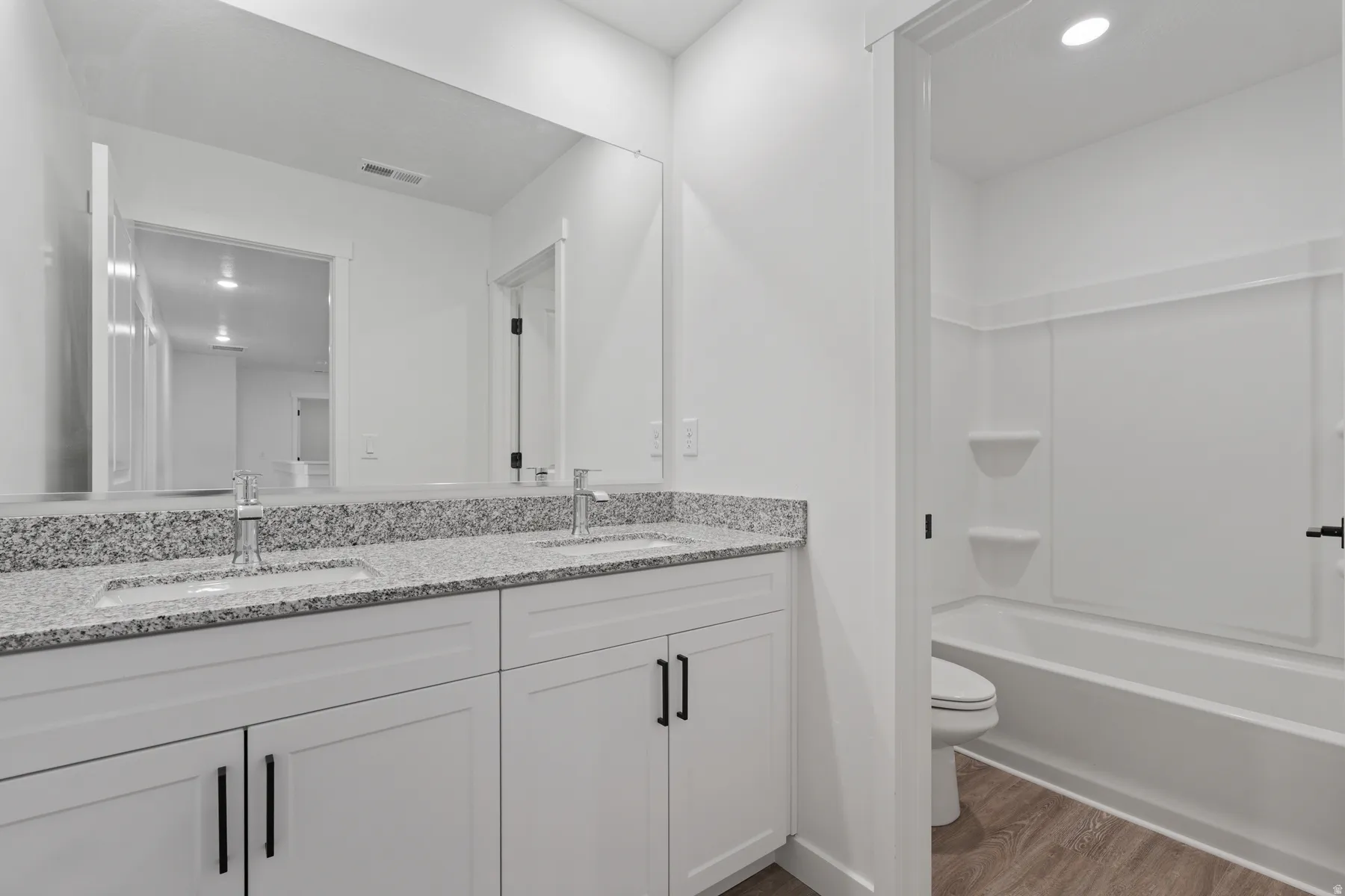 Double vanity with granite-style countertop, white cabinetry with black hardware, and wood-finish flooring