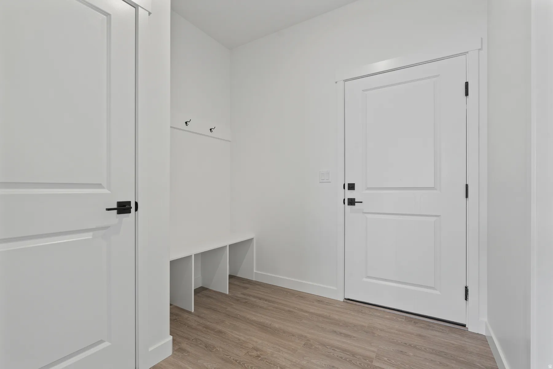 Built-in bench with storage cubbies and wall-mounted hooks, wood-finish flooring, and white paneled doors with matte black hardware