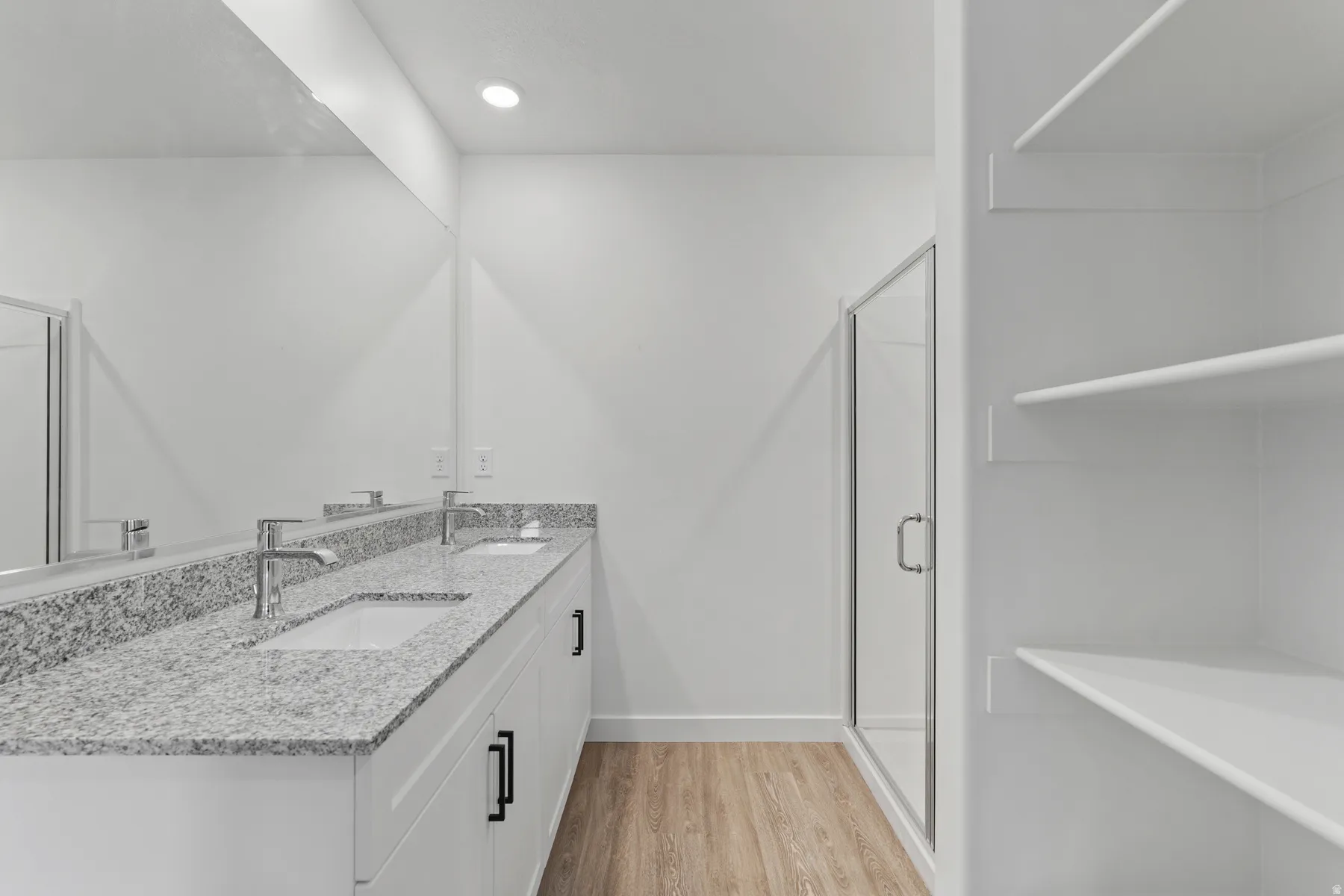 Dual vanity with undermount sinks, granite countertop, built-in shelving, glass-enclosed shower, and wood-finish flooring