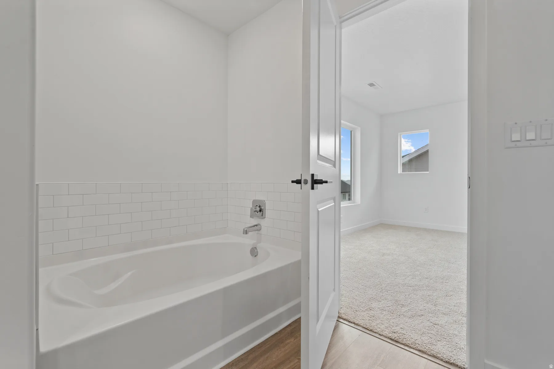 White subway tile surround with chrome fixtures, wood-finish flooring, white paneled door with matte black hardware, and two windows