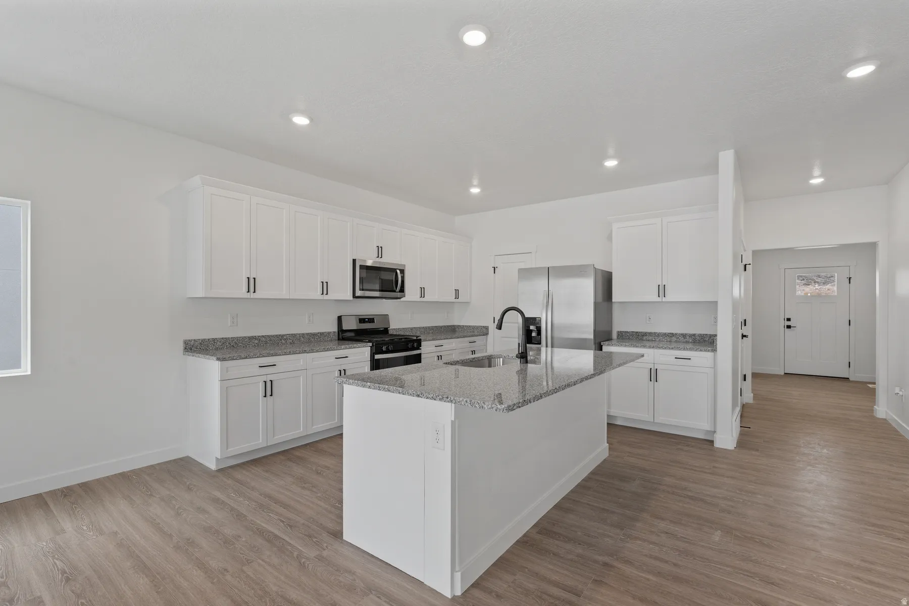 Modern kitchen featuring white cabinetry, stone-finish countertops, an island with an integrated sink, stainless steel appliances, and wood-finish flooring