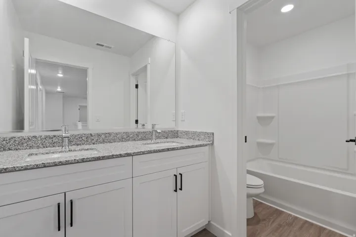 Double vanity with granite-style countertop, white cabinetry with black hardware, and wood-finish flooring