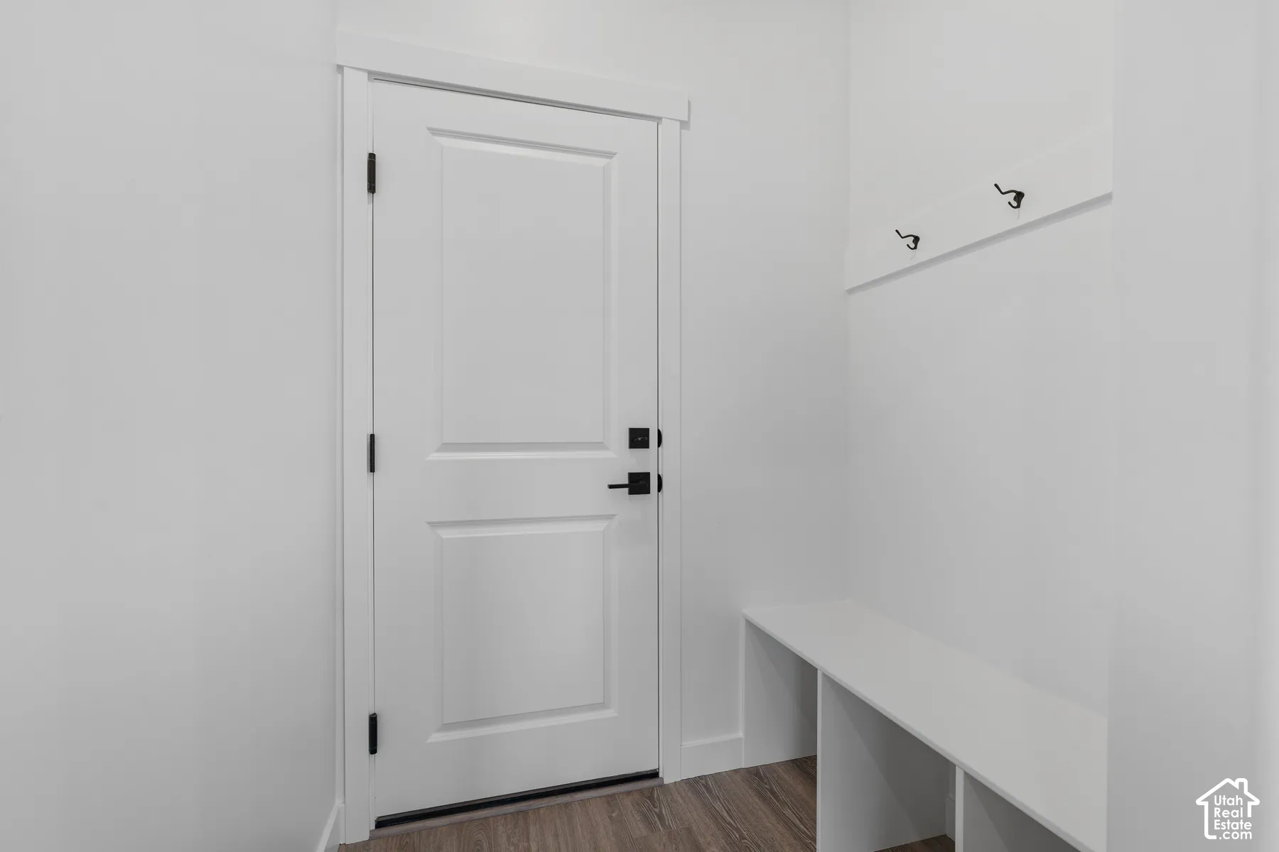 White paneled door with matte black hardware, wood-finish flooring, built-in storage bench, and wall-mounted coat rack