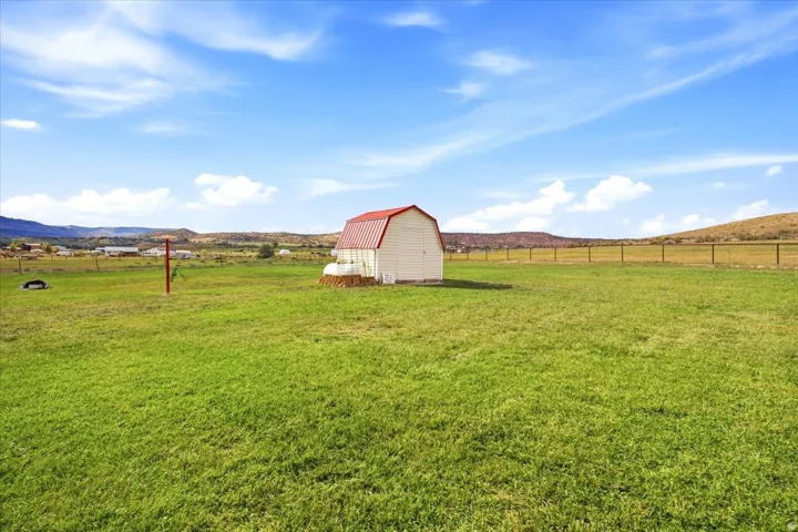 Expansive grassy acreage featuring a white outbuilding with a red gable roof, a perimeter fence, and distant rolling hills