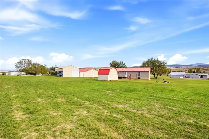 Expansive green lawn with distant mountain backdrop, featuring a primary residence with a red metal roof, a detached garage with a white overhead door, an additional red-roofed outbuilding, and a small red shed
