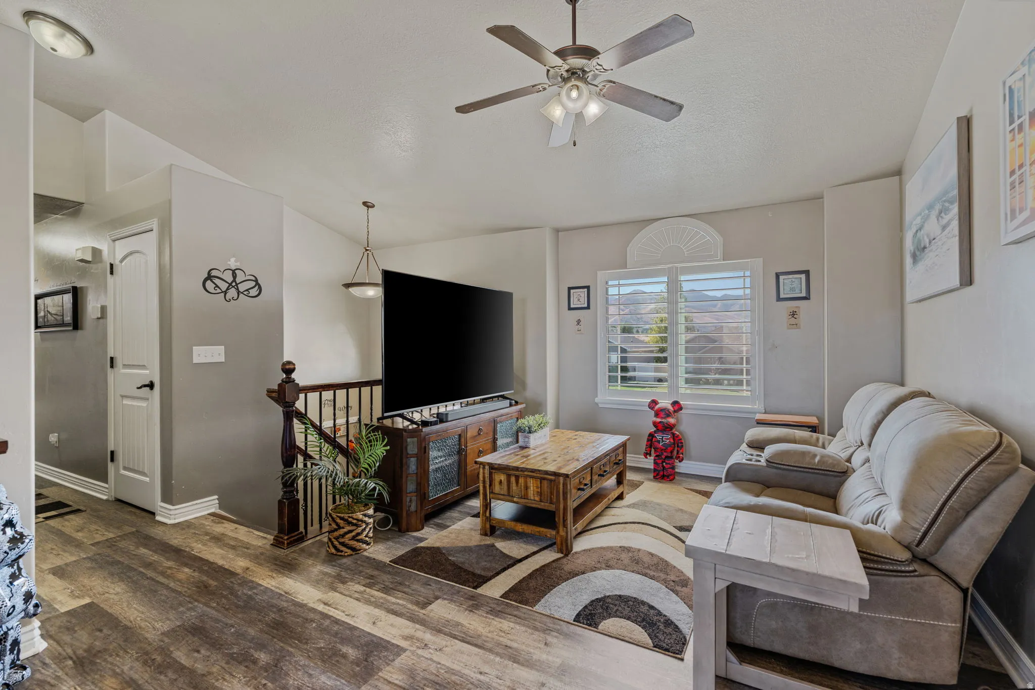 Wood-finish flooring, a ceiling fan with integrated lighting, a window with plantation shutters, a decorative arched transom window, and a built-in wooden media console