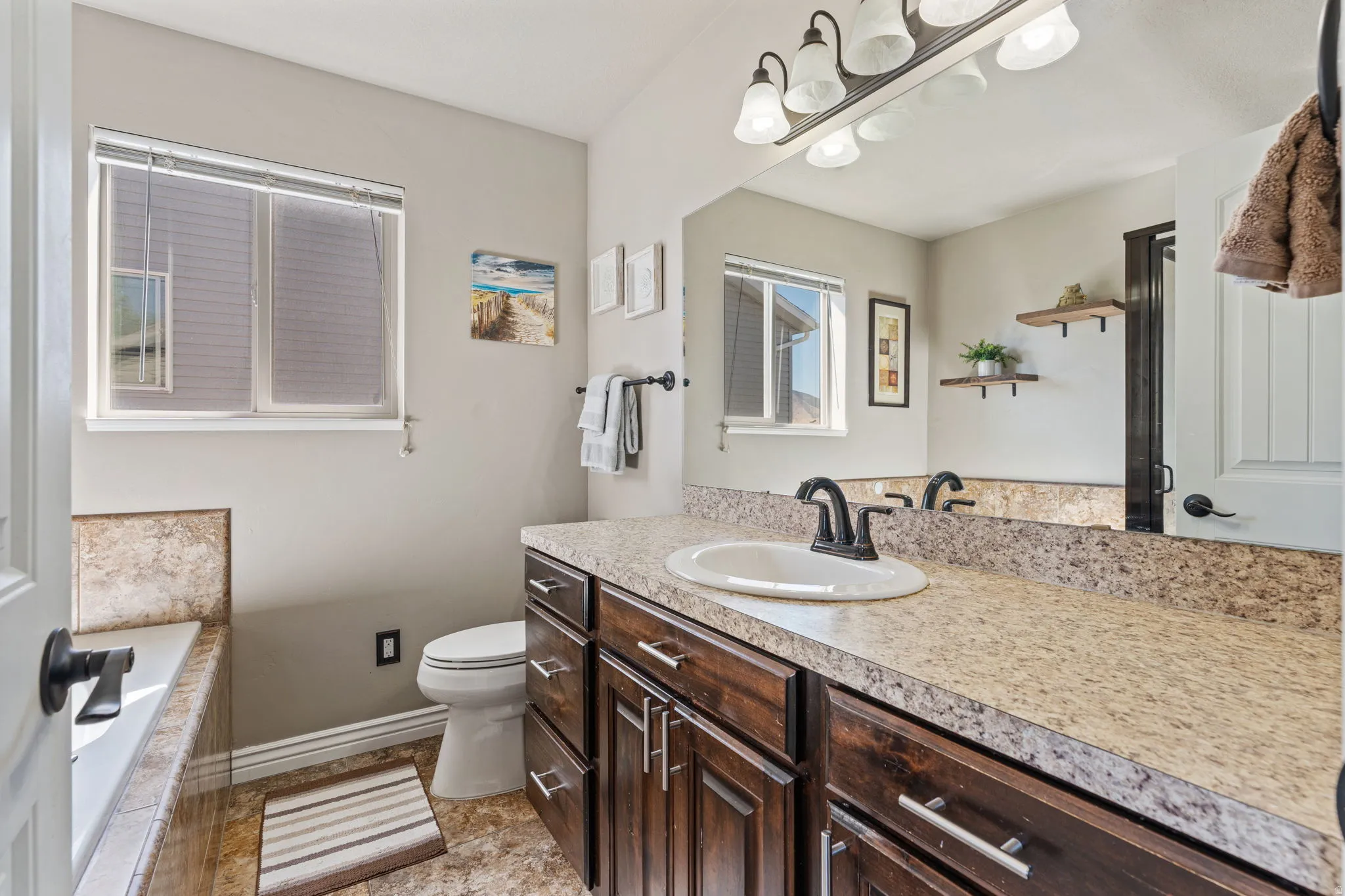 Bathroom featuring a built-in tub with tile surround, a single vanity with an integrated sink and dark wood-finish cabinetry, a large wall mirror, and a multi-light fixture