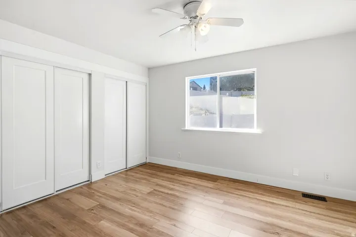 Wood-finish flooring, white sliding closet doors, white walls, ceiling fan with light, and a window providing natural light