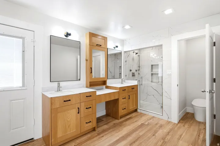 Dual wood-finish vanities with integrated sinks and light countertops, a separate makeup vanity, a glass-enclosed shower with tile surround, and wood-finish flooring