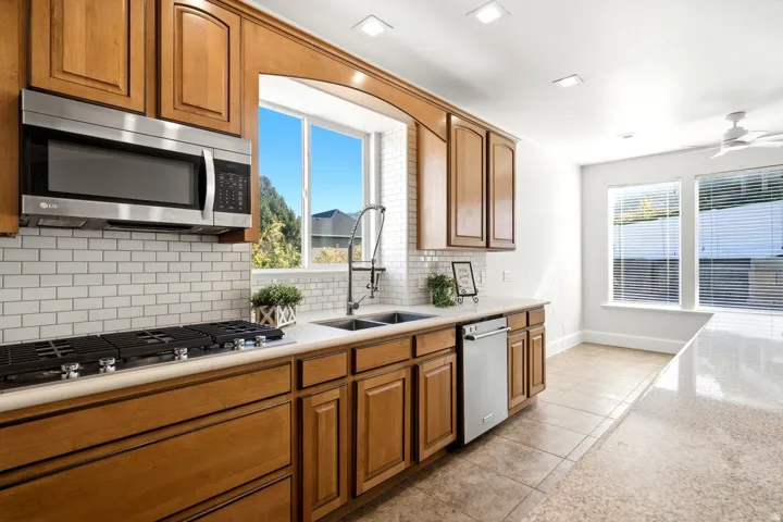 Spacious kitchen featuring warm wood cabinetry, stainless steel appliances including a gas cooktop and microwave, subway tile backsplash, and a large window overlooking the grounds