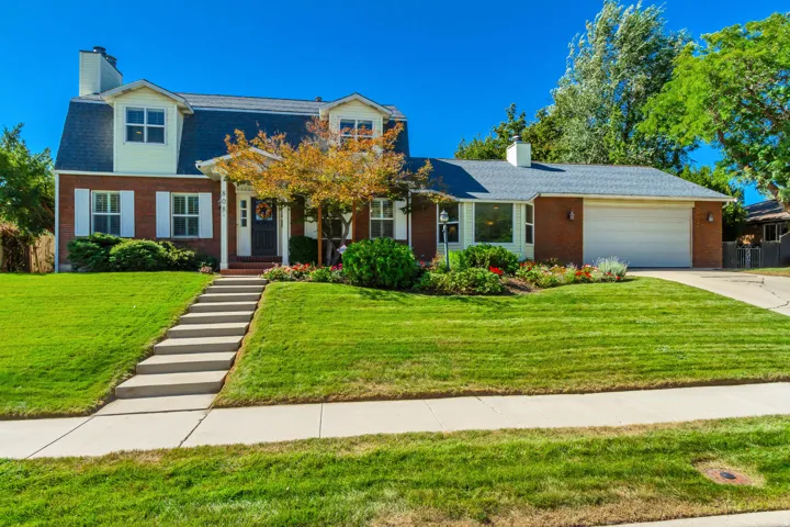 Brick facade with white shutters, dormer windows, attached garage, concrete steps, and a landscaped front yard