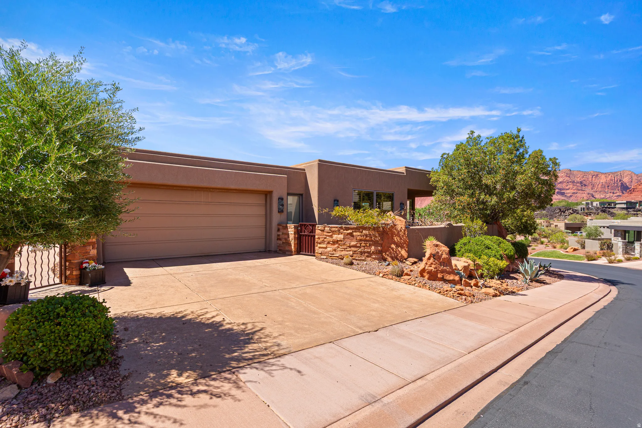 Stucco exterior with an attached garage, a private gated entry, and desert landscaping featuring natural rock elements and mature trees