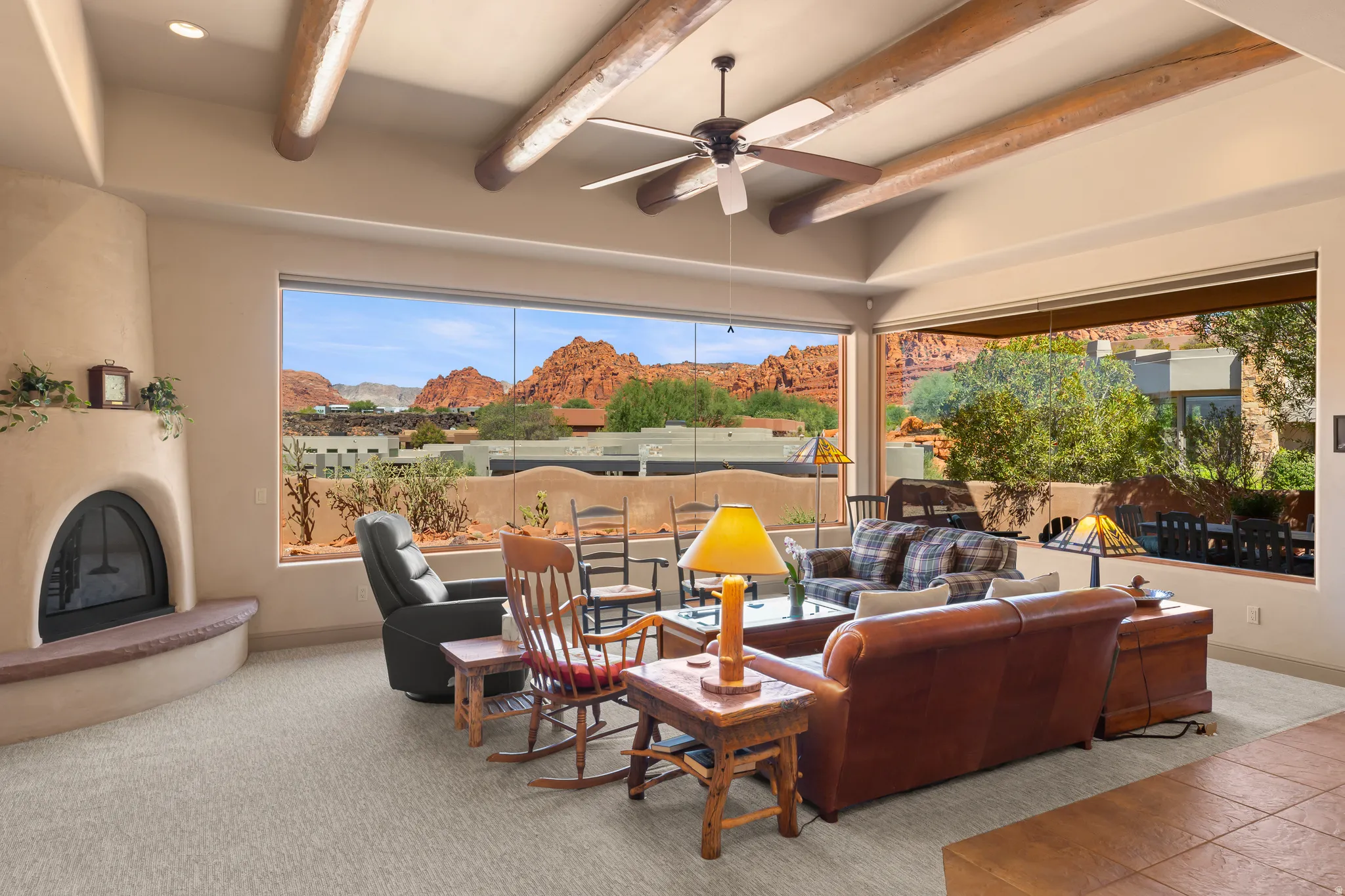 Exposed wood beams, ceiling fan, expansive windows, stucco fireplace, and surrounding desert mountain scenery