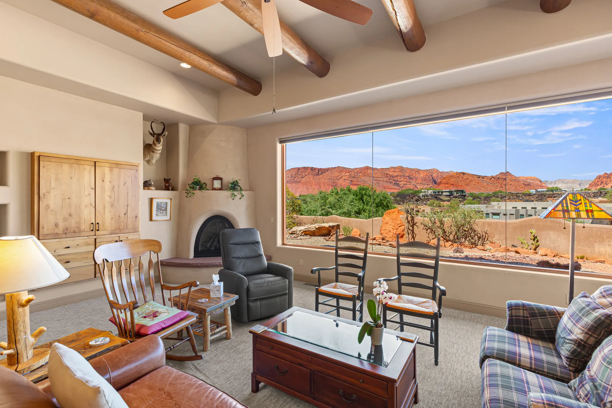 Spacious living area featuring exposed ceiling beams, a built-in entertainment center, a kiva fireplace with a hearth, and expansive floor-to-ceiling windows showcasing a red rock landscape