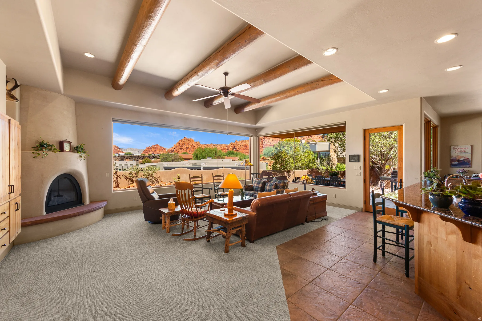 Expansive window with mountain scenery, exposed ceiling beams, recessed lighting, built-in adobe-style fireplace, and an integrated kitchen island with a stone-finish countertop