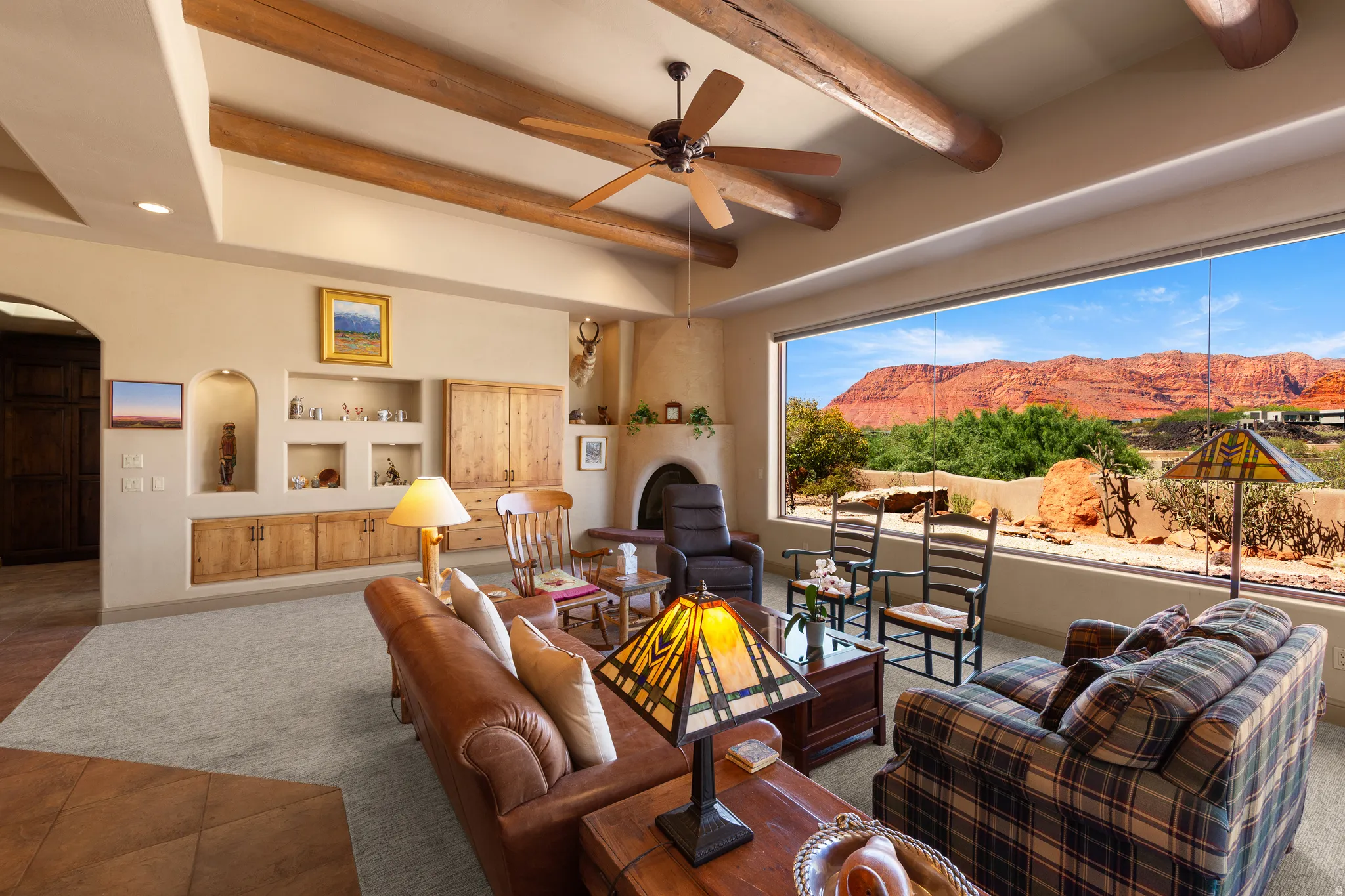 Expansive window overlooking rugged desert landscape, exposed wood beam ceiling, built-in wall niches, wood-finish cabinetry, and a corner fireplace