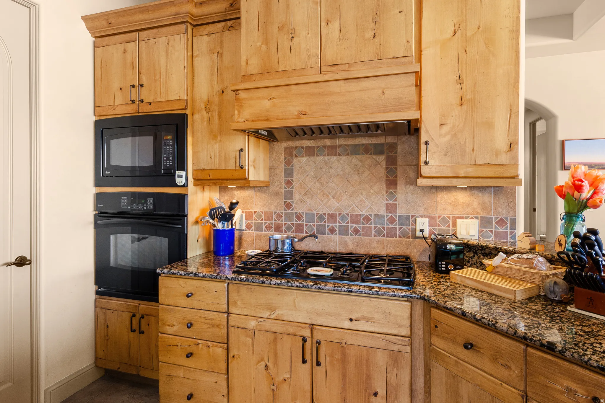 Built-in black microwave and oven, gas cooktop, natural wood cabinetry, granite countertops, and decorative tile backsplash
