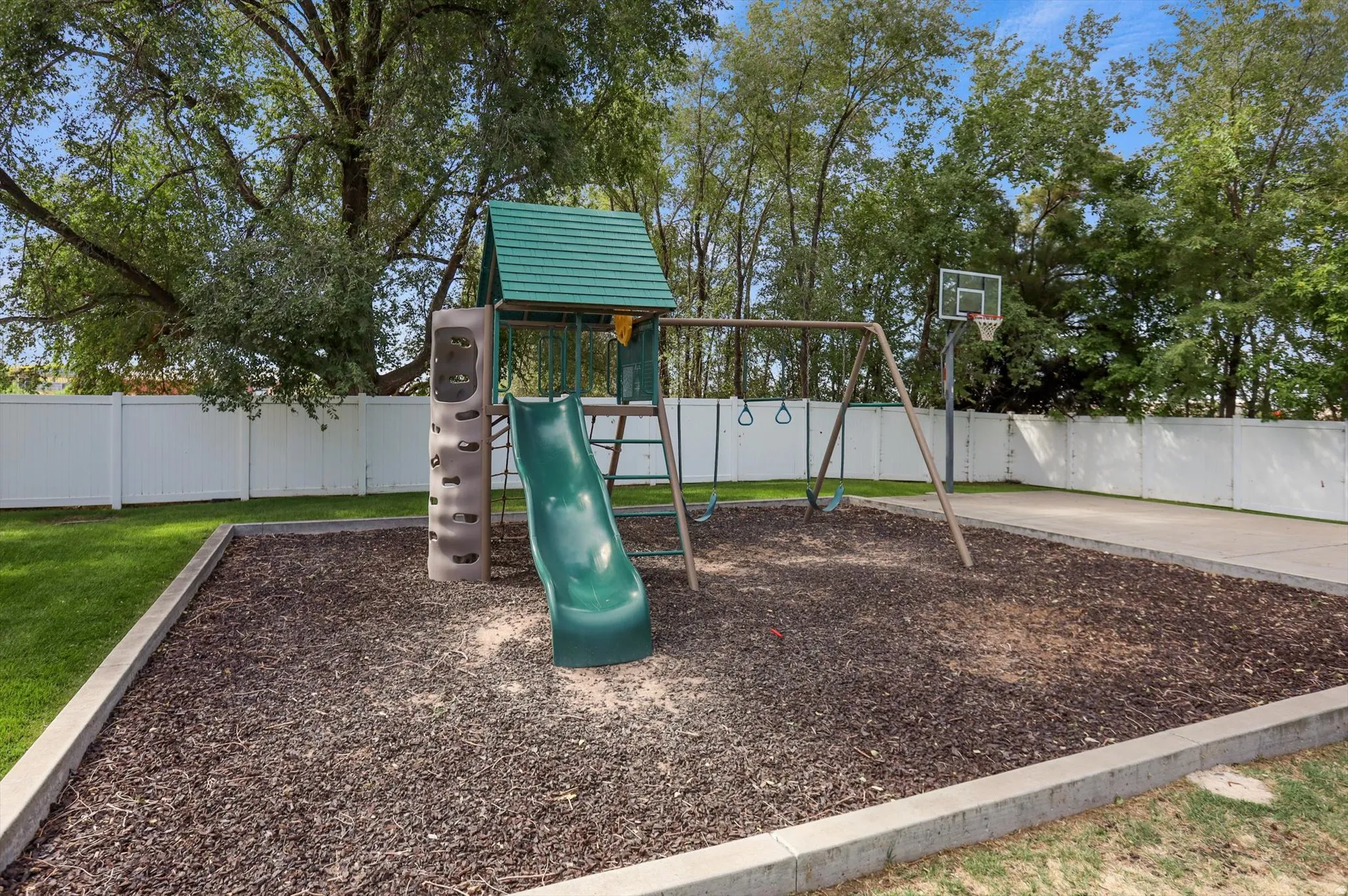 Outdoor play structure with slide and swings, basketball hoop, and white privacy fencing