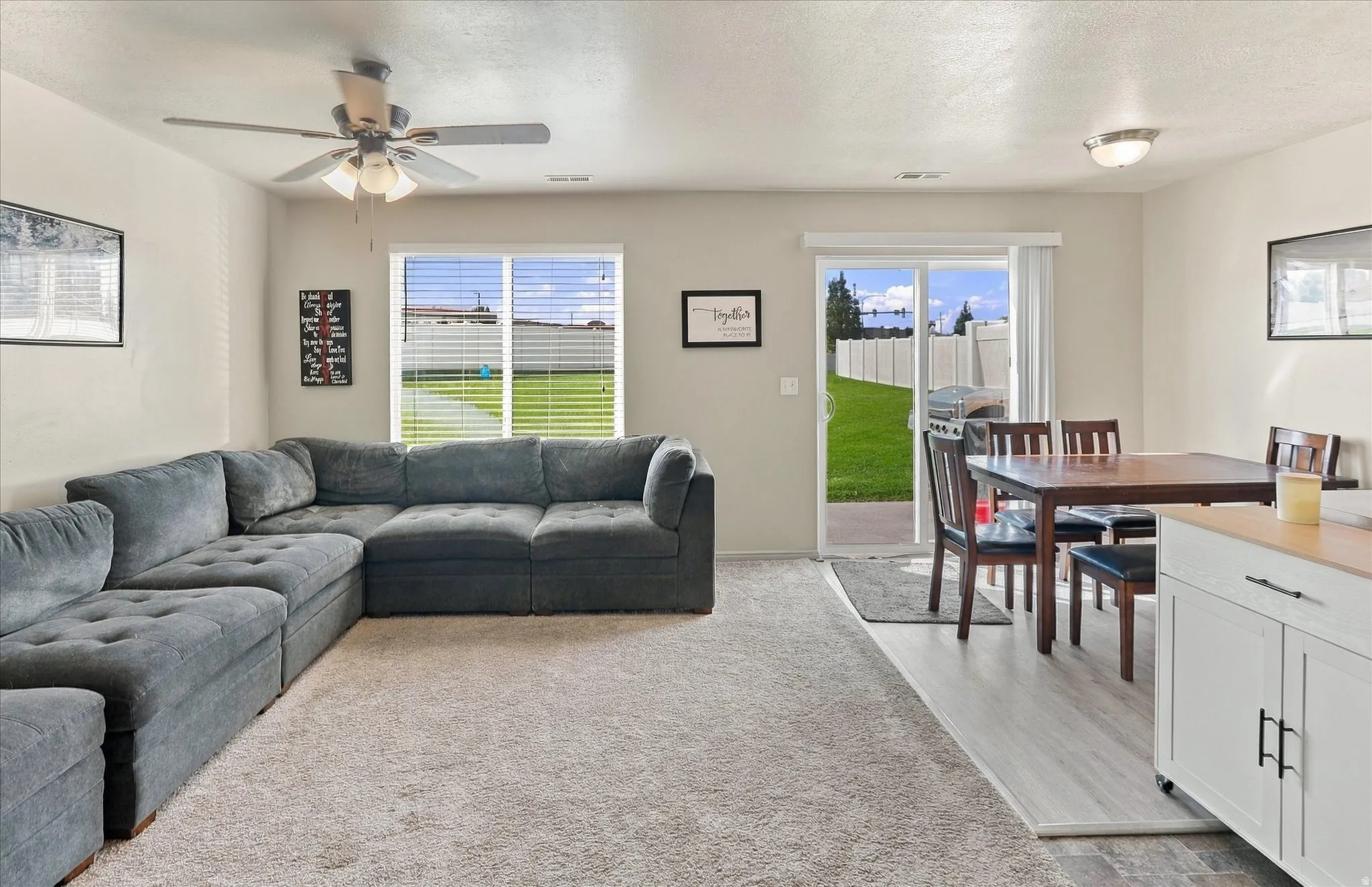 Carpeted living area with a ceiling fan, dual windows, and a sliding glass door opening to a private fenced yard