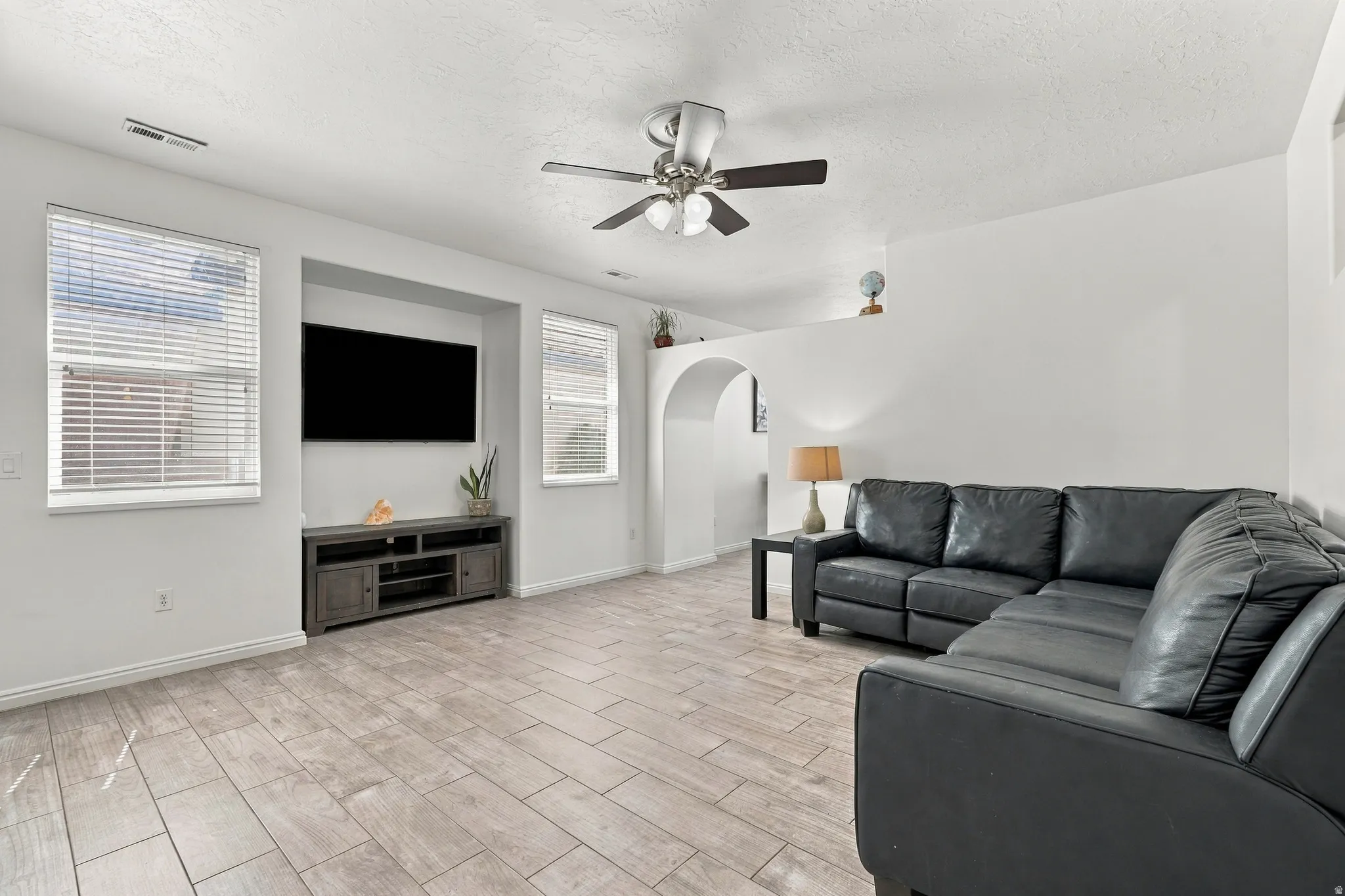 Wood-finish tile flooring, recessed media niche, multiple windows with blinds, ceiling fan with light, and arched doorway