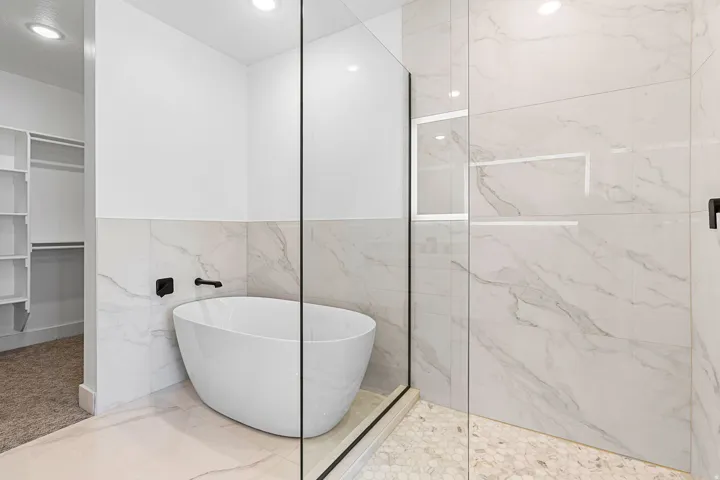 Freestanding soaking tub with matte black fixtures, glass-enclosed shower with a recessed shelf, and large-format tile walls