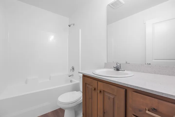 Wood-finish flooring, white bathtub with shower, white toilet, wood vanity with an integrated sink, and a speckled countertop