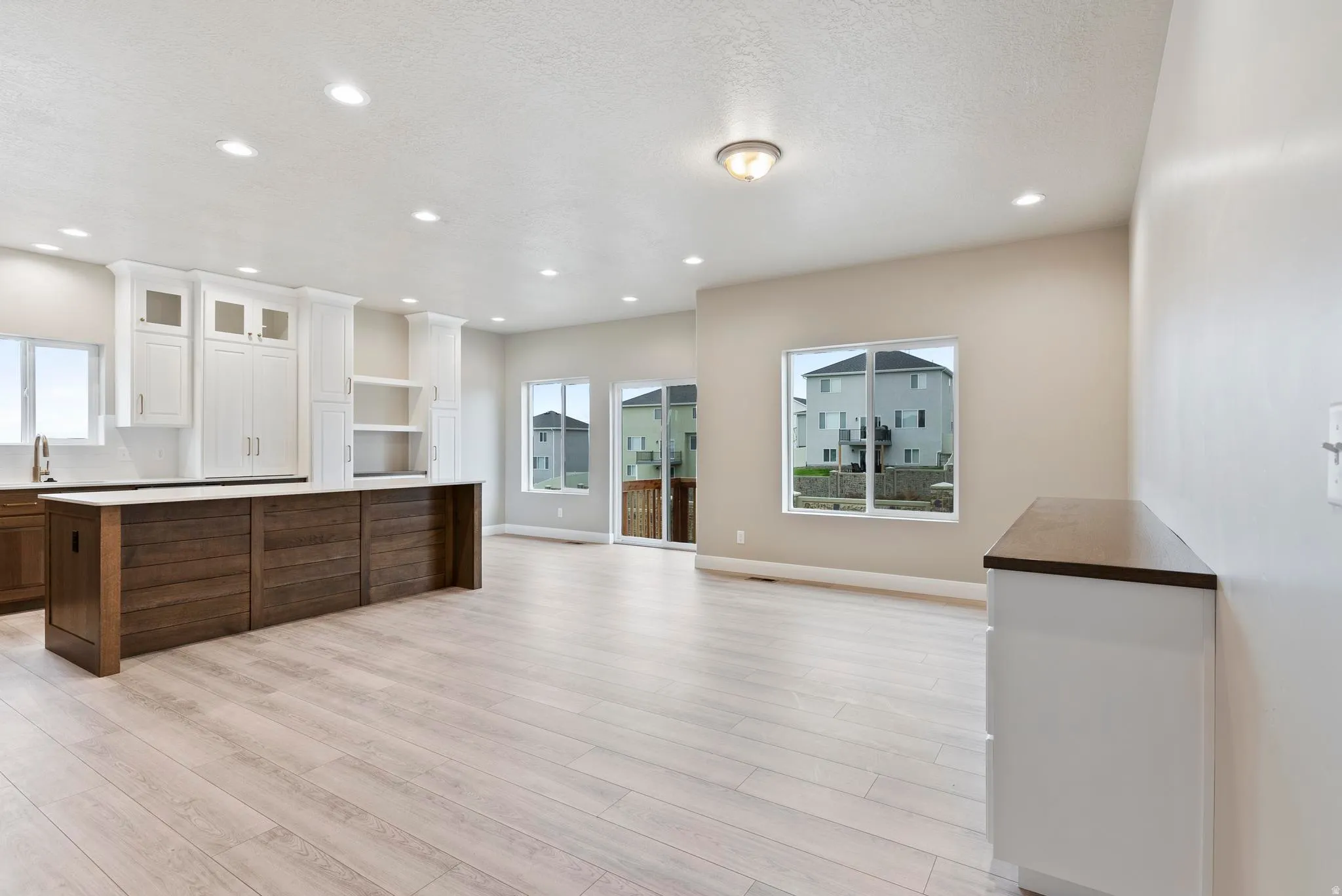 Open-concept living area with light wood-finish flooring, recessed lighting, and a kitchen island featuring a wood-panel base and light-toned countertop