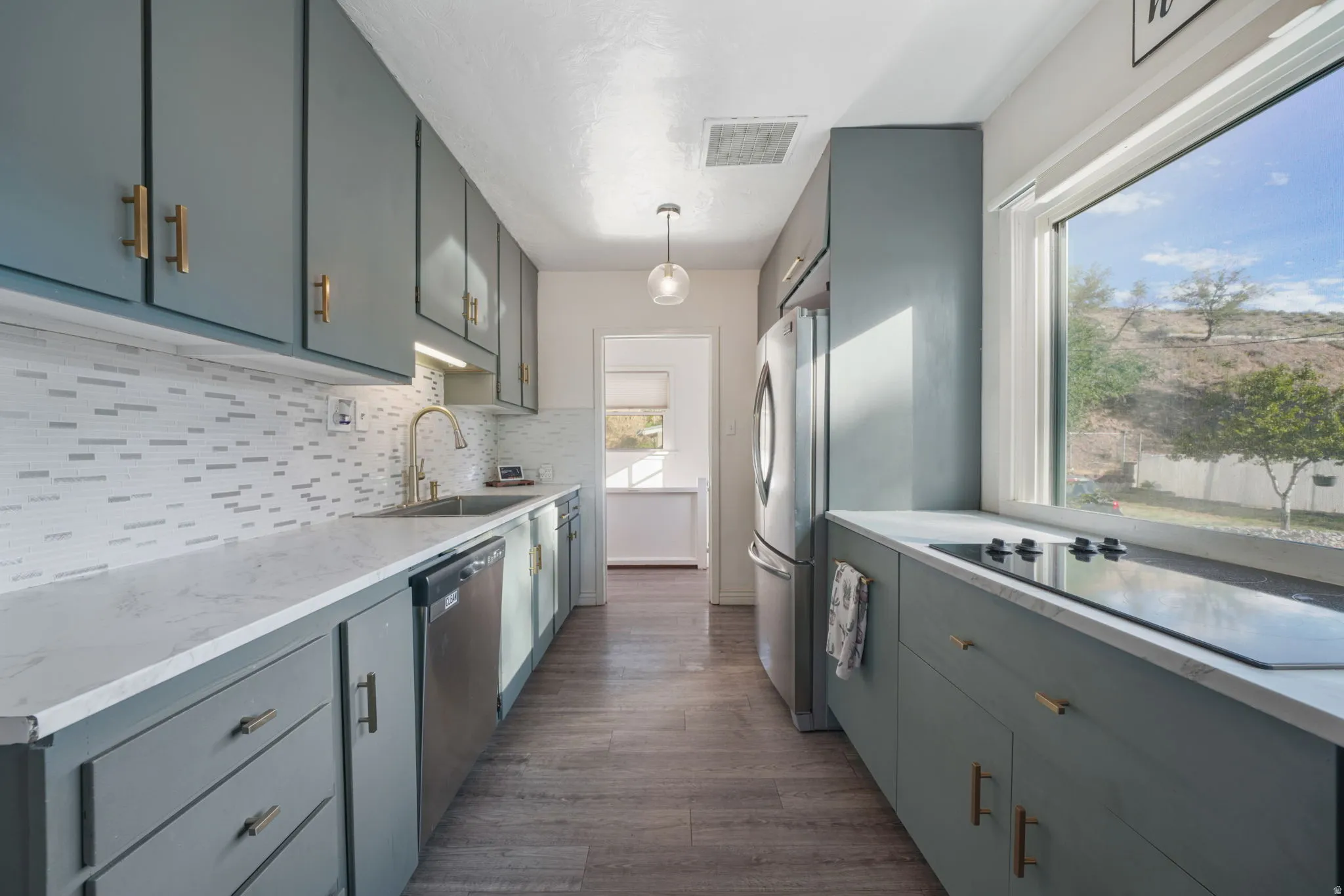 Galley kitchen featuring matte blue cabinetry with gold-toned hardware, light-toned countertops, tile backsplash, and stainless steel appliances