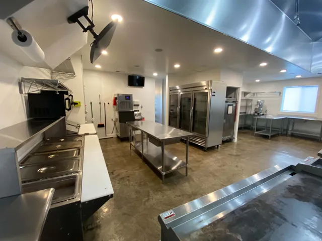 Commercial kitchen featuring stainless steel work surfaces, recessed lighting, concrete flooring, large refrigeration units, and a window