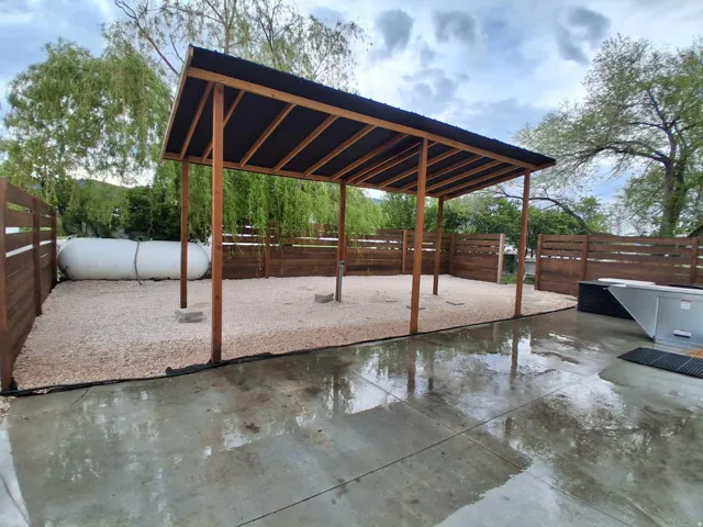 Covered outdoor structure with wood posts and a dark roof, featuring a gravel ground surface, wood fencing, and a concrete patio