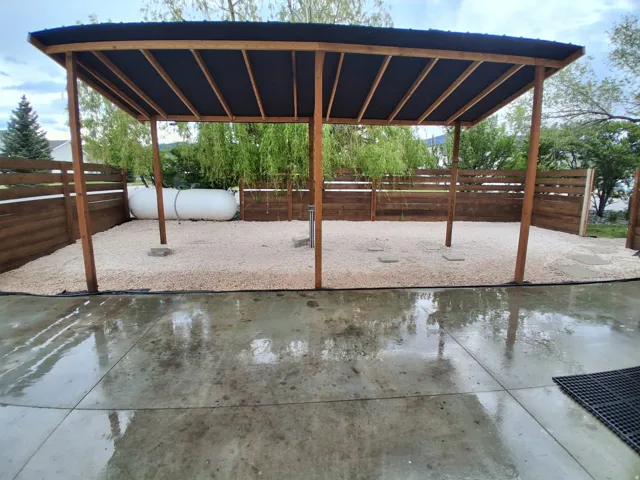 Outdoor patio area featuring a covered pergola with a dark roof and exposed wood framing, a gravel-covered ground surface, and a horizontal wood fence