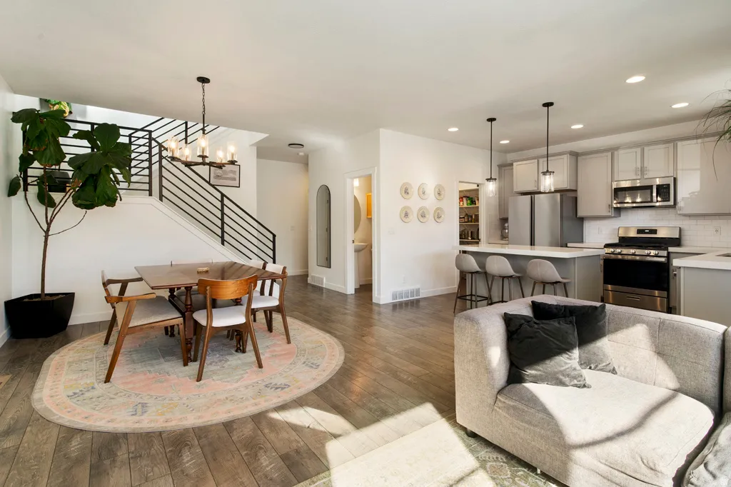 Open-concept living space with wood-finish flooring, a modern kitchen featuring stainless steel appliances and light cabinetry, a dining area with a contemporary chandelier, and a staircase with sleek black metal railings
