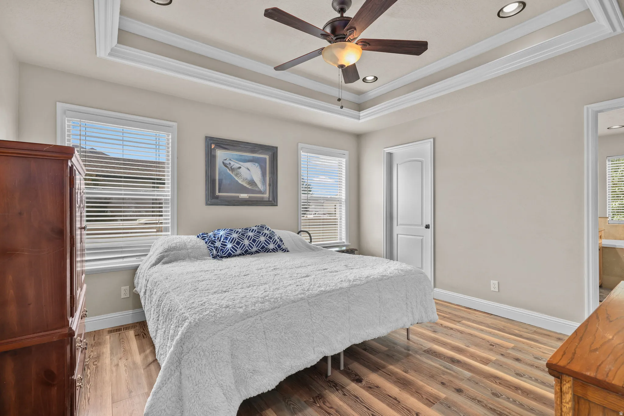 Tray ceiling with crown molding, ceiling fan with integrated lighting, recessed lighting, wood-finish flooring, and white window blinds