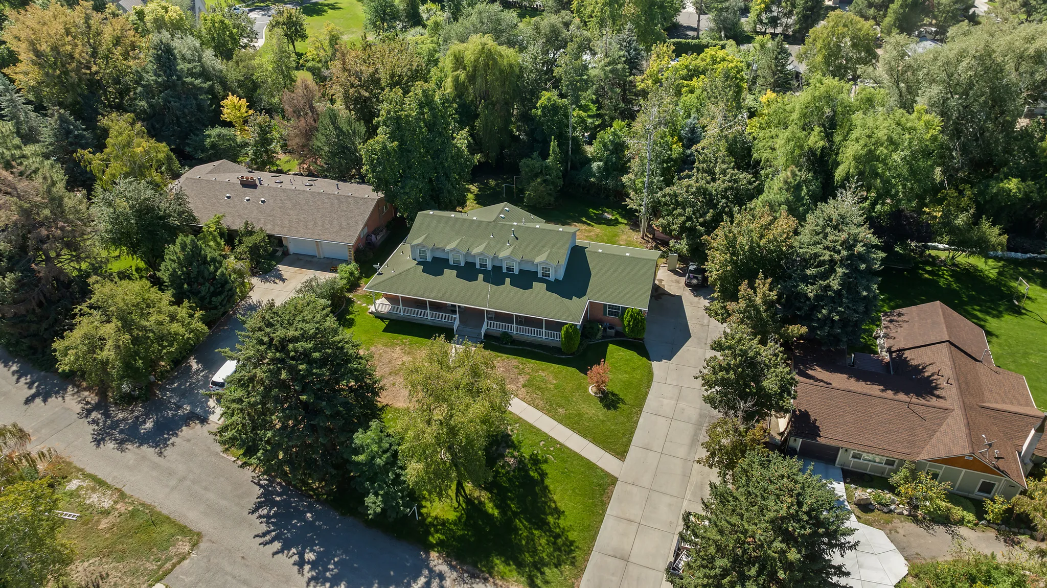 Green roof with multiple dormer windows, covered front porch, mature trees, and a concrete driveway