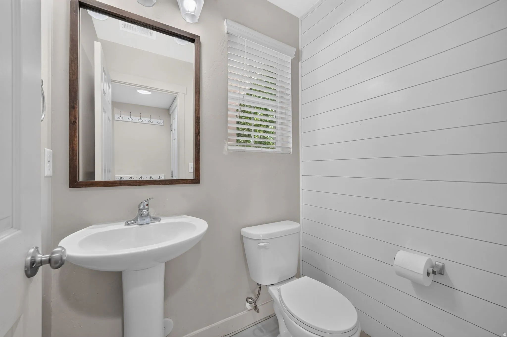 Pedestal sink with chrome faucet, framed mirror, wall-mounted light fixture, white horizontal paneling, and window with white blinds