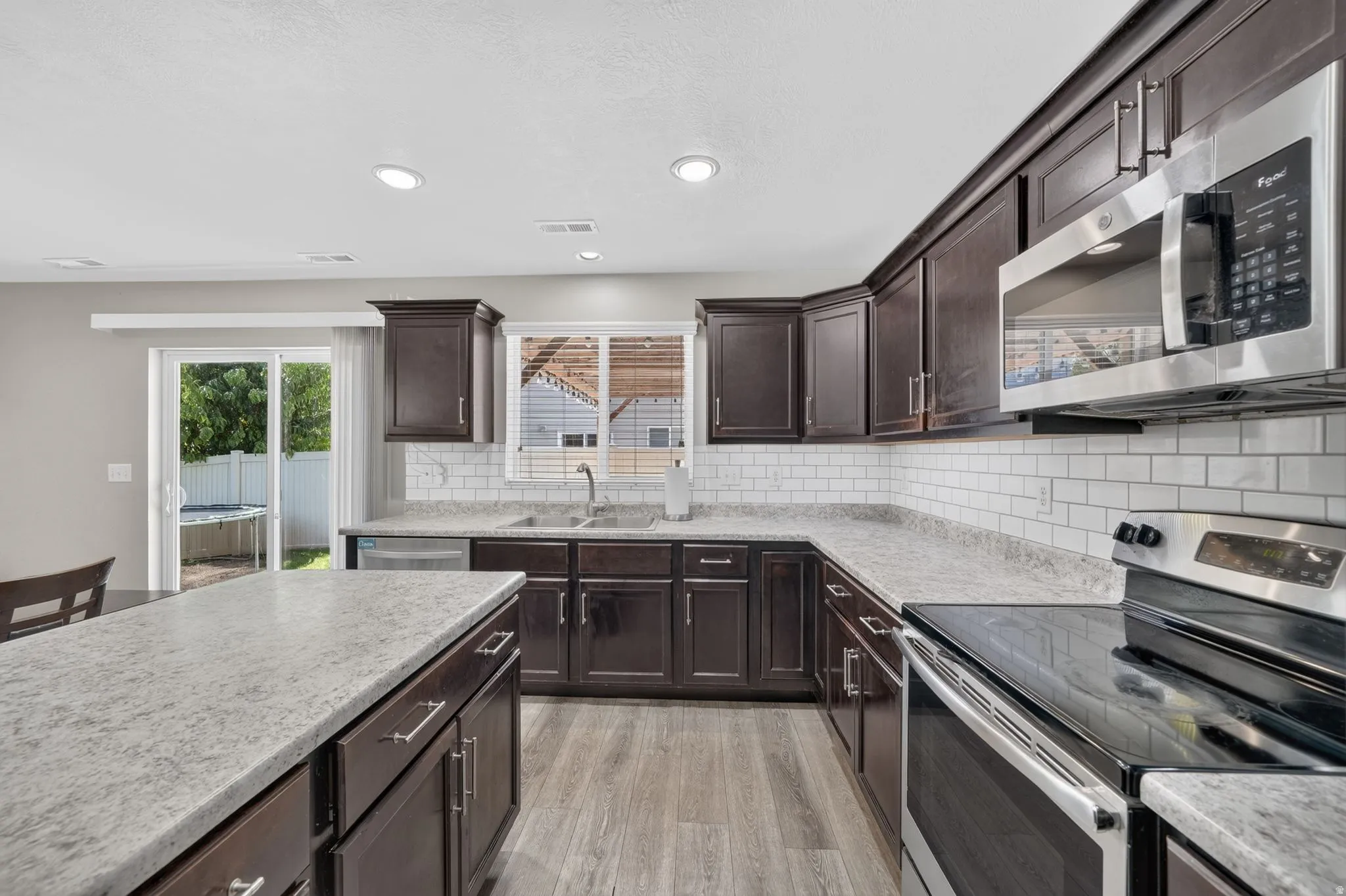 Dark wood-finish cabinetry with silver hardware, light-toned countertops, white subway tile backsplash, stainless steel appliances, and wood-finish flooring