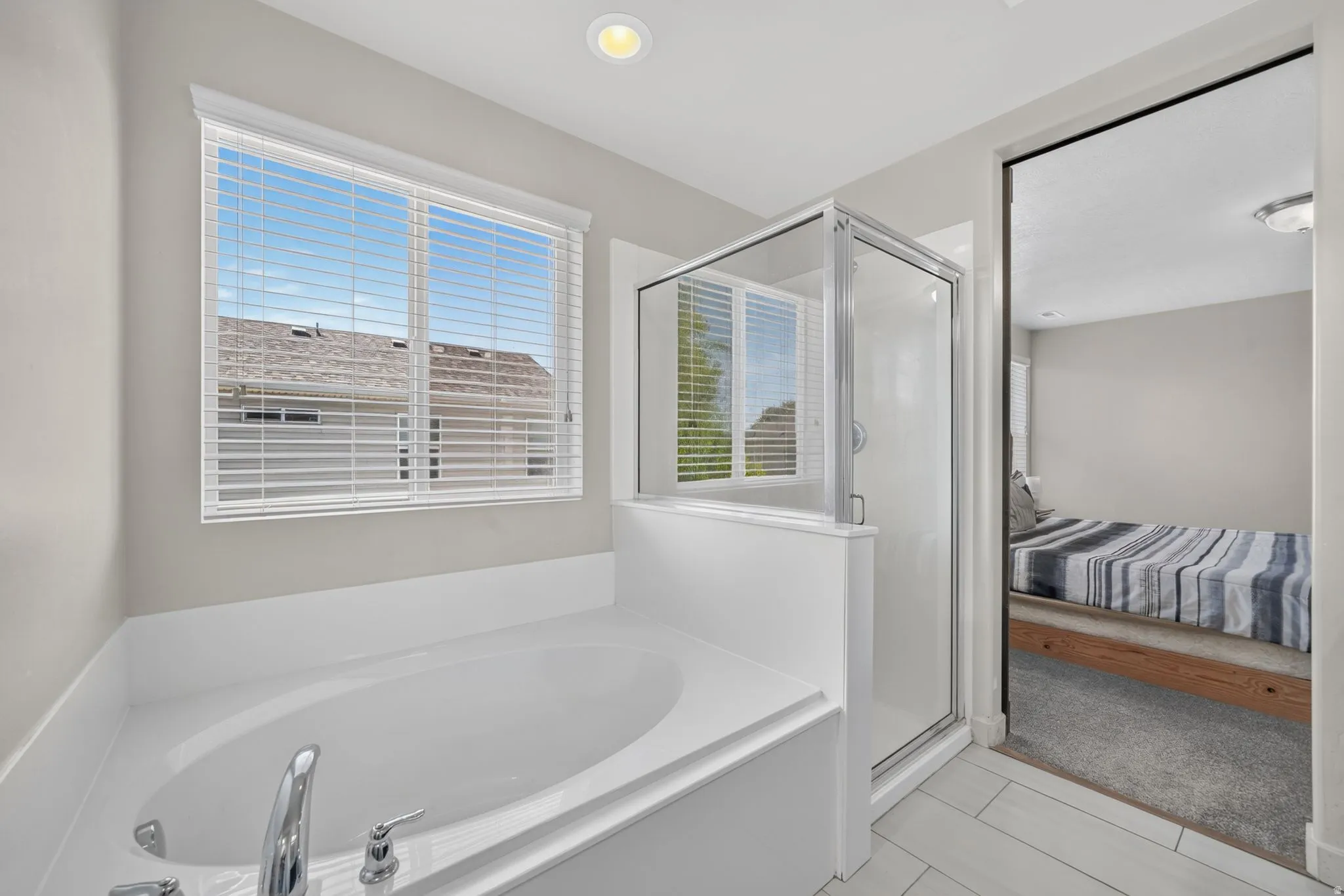 Built-in soaking tub with chrome fixtures, glass-enclosed shower, white tile flooring, and a window with blinds
