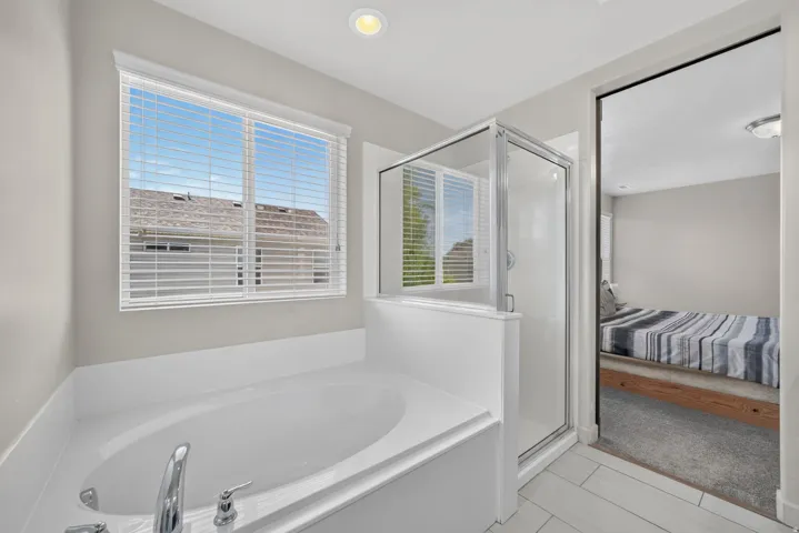 Built-in soaking tub with chrome fixtures, glass-enclosed shower, white tile flooring, and a window with blinds