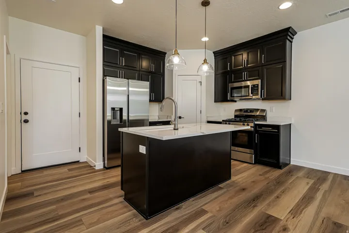 Kitchen featuring a central island with a sink, black cabinetry, stainless steel appliances, pendant lighting, and wood-finish flooring