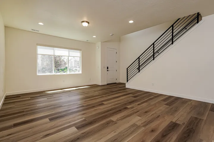 Spacious living area featuring wood-finish flooring, a large window with roller shades, recessed lighting, and a contemporary black metal stair railing