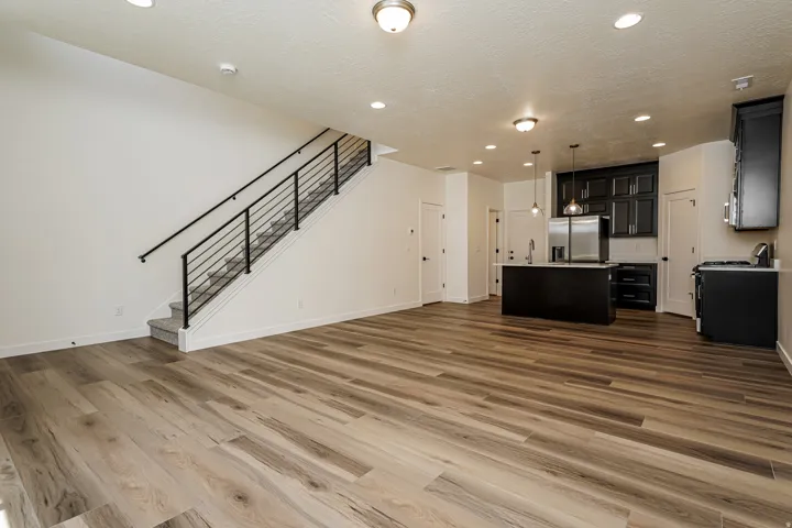 Spacious open-concept living area featuring wood-finish flooring, a modern kitchen with dark cabinetry, a kitchen island with pendant lighting, stainless steel appliances, and a carpeted staircase with contemporary metal railings