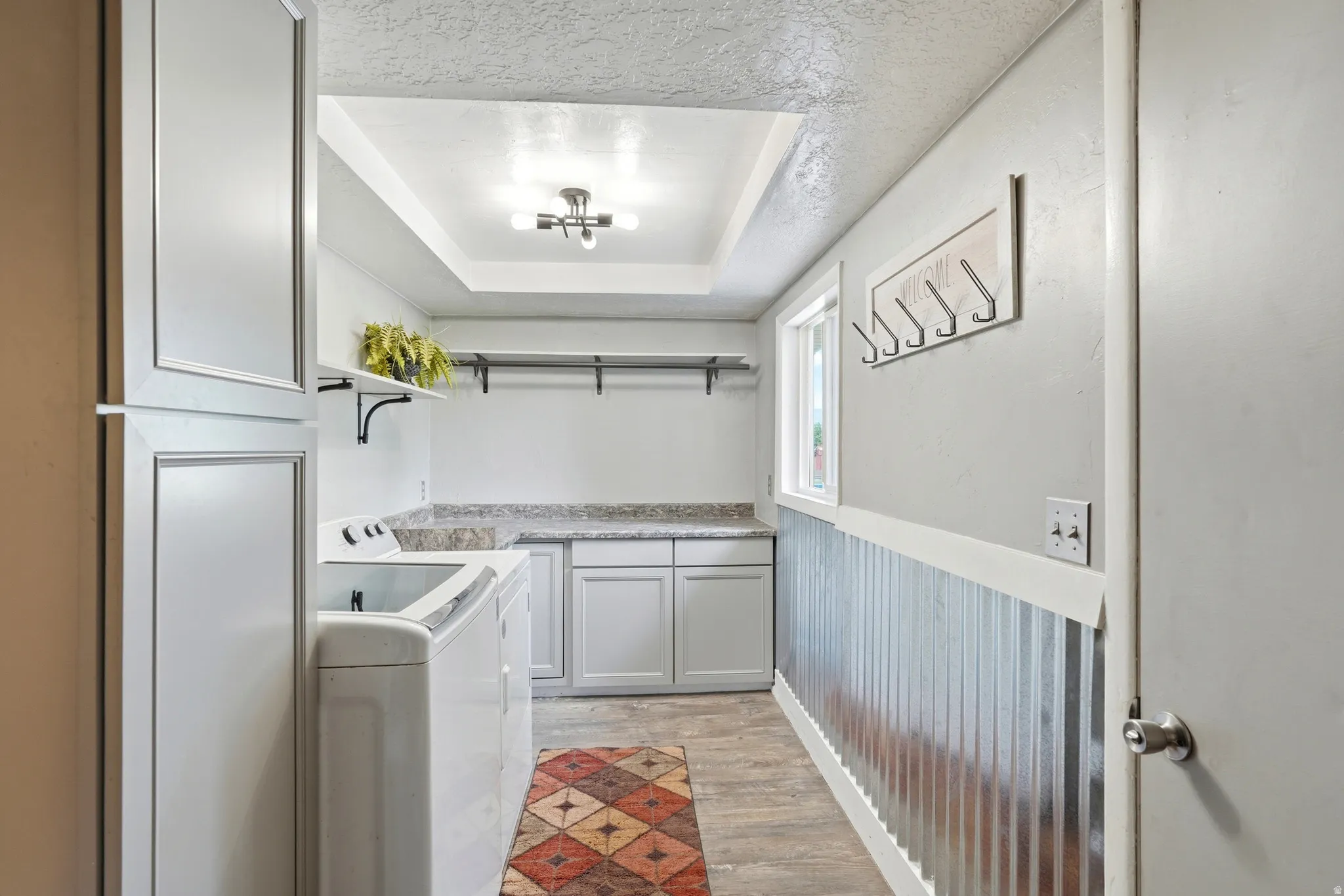 Laundry area featuring upper and lower cabinetry, a tray ceiling, wood-finish flooring, a window, and a corrugated metal wainscoting
