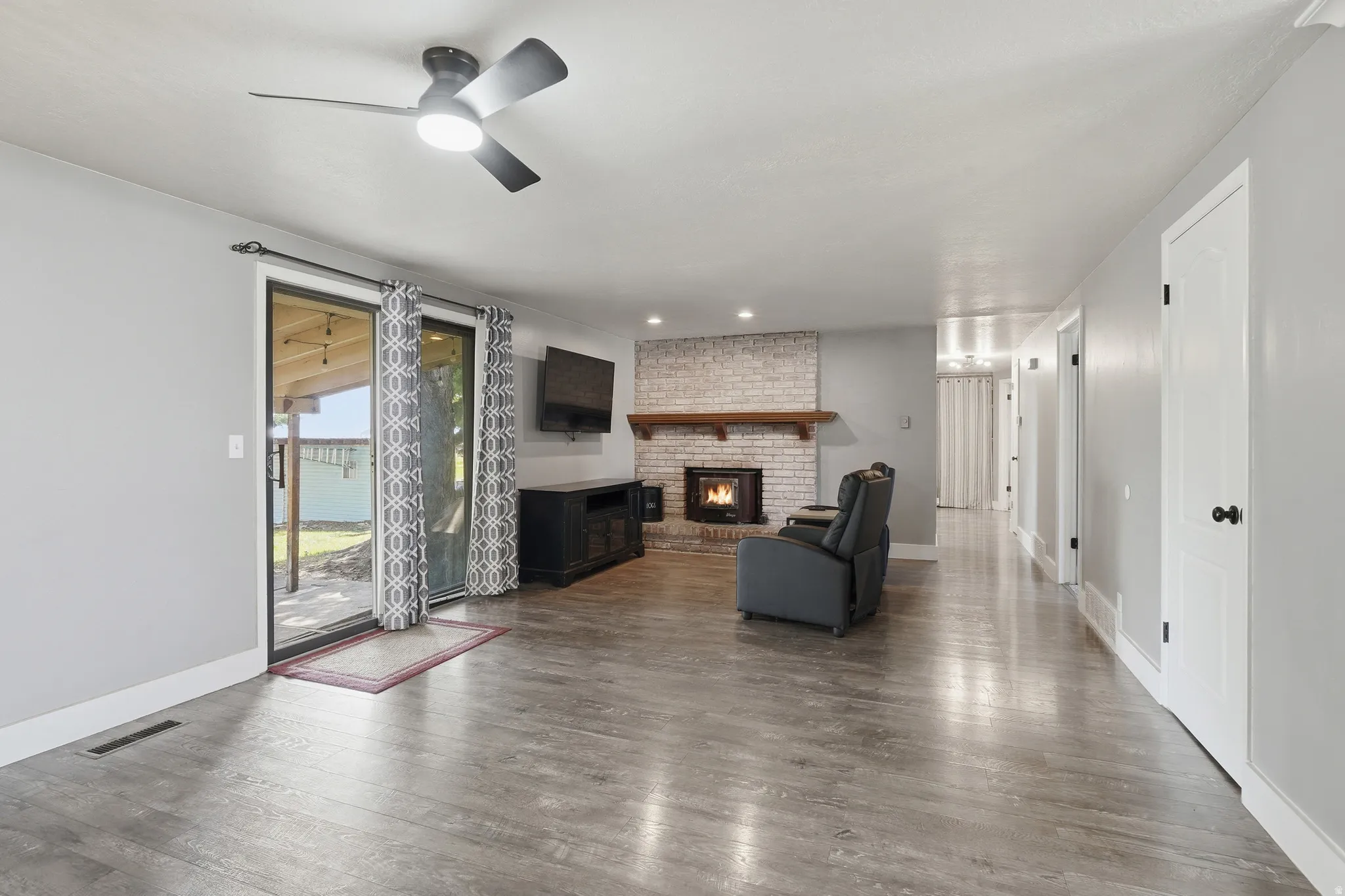 Wood-finish flooring, brick fireplace with wood mantel, sliding glass patio door, ceiling fan with light, and light gray wall paint