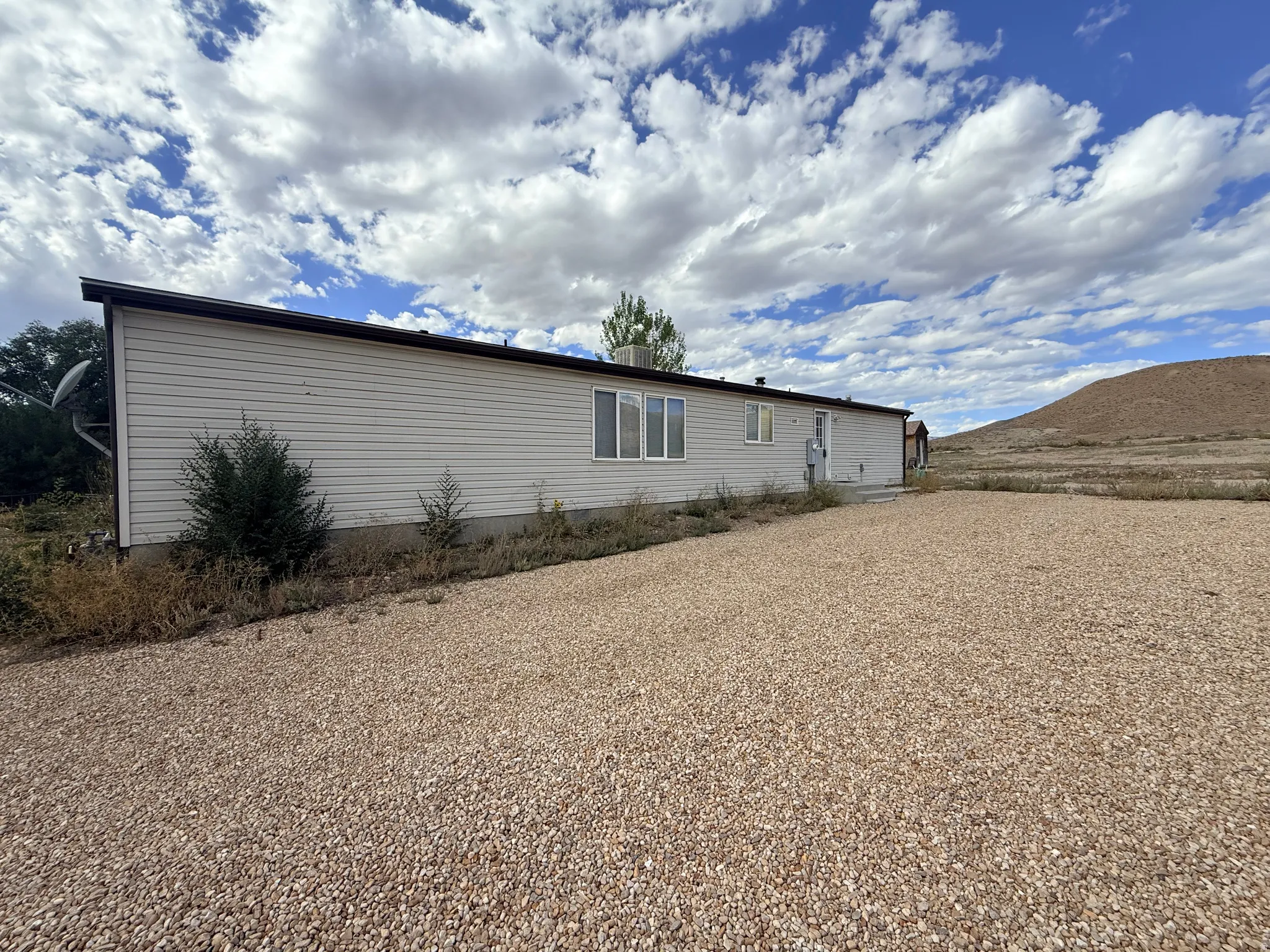 Gravel driveway extending along the side of the residence with horizontal siding, multiple windows, and a partially visible distant hill