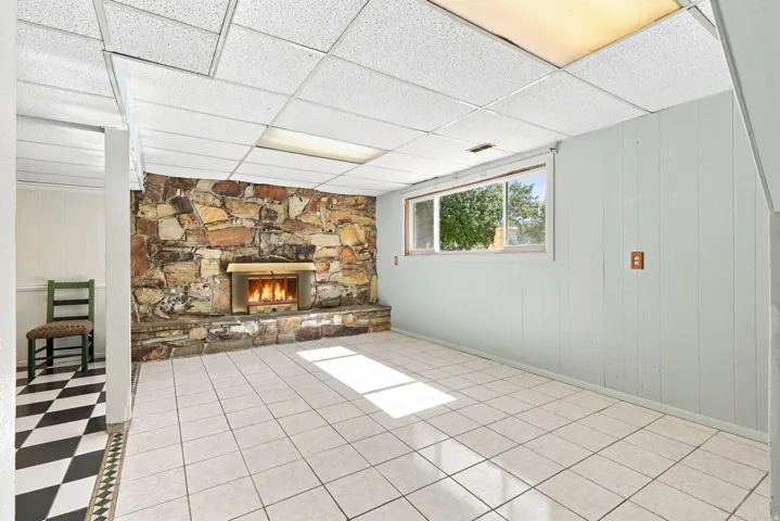 Stone-clad fireplace with a hearth, white tile flooring, drop ceiling with integrated lighting, and a casement window