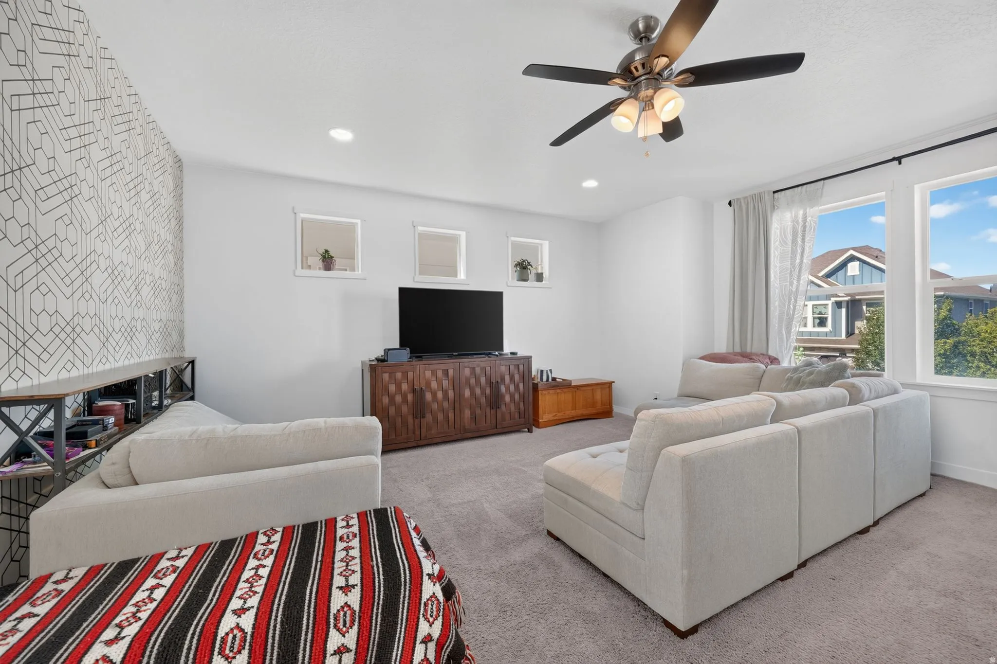 Carpeted living space featuring recessed lighting, a ceiling fan with integrated light fixture, a geometric accent wall, and multiple windows