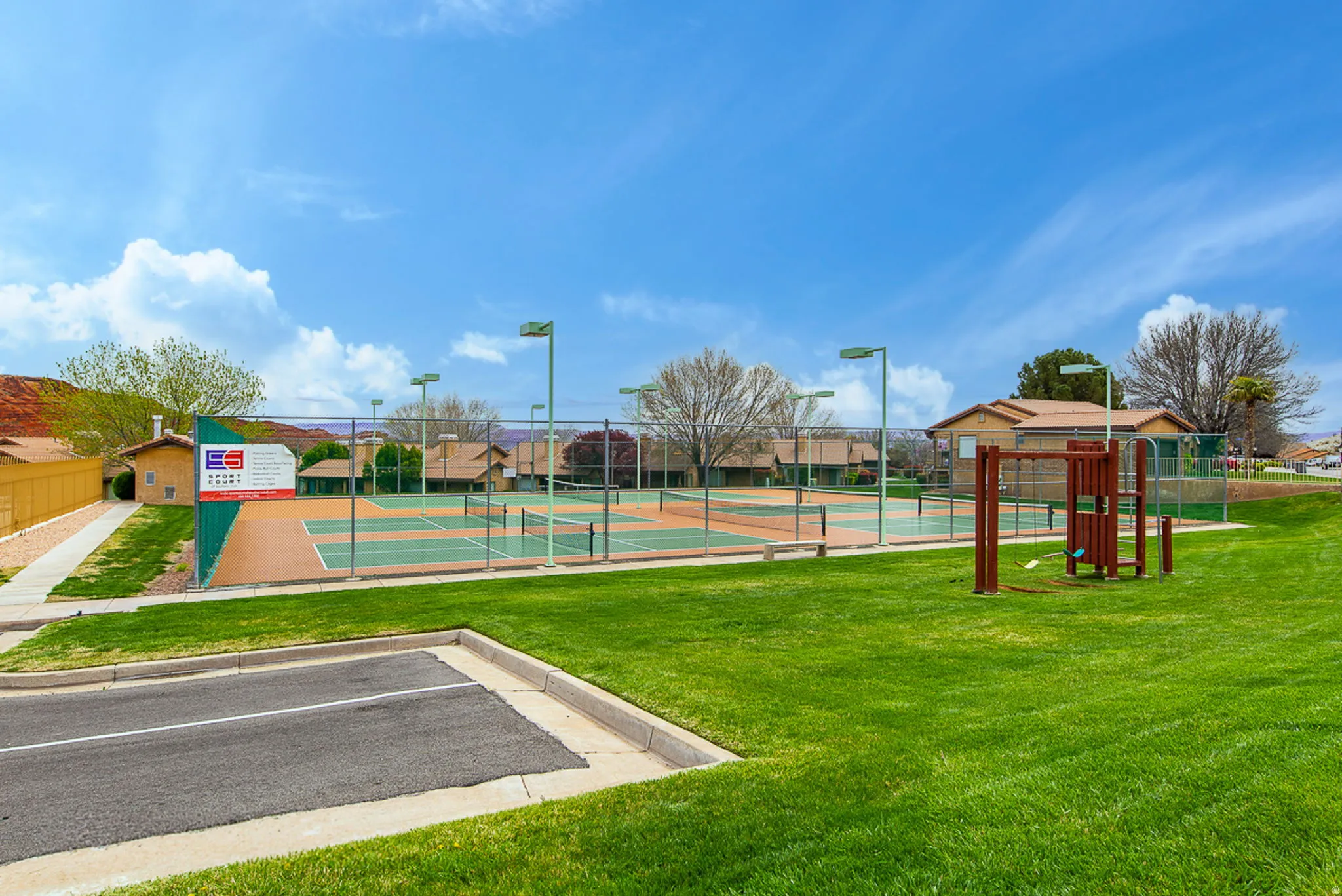 Expansive green space featuring multiple tennis courts, a playground structure, and overhead lighting