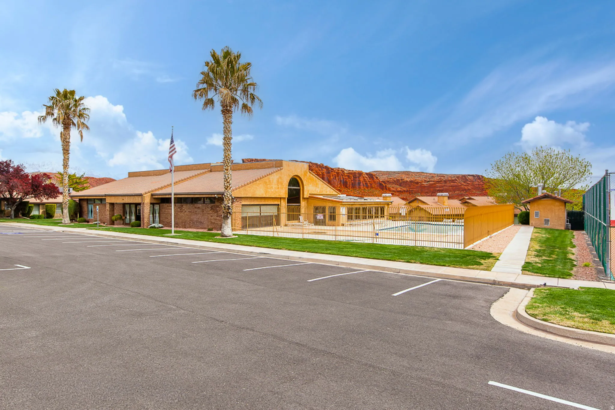 Expansive parking area, stucco exterior, multiple palm trees, fenced outdoor pool, and tennis court