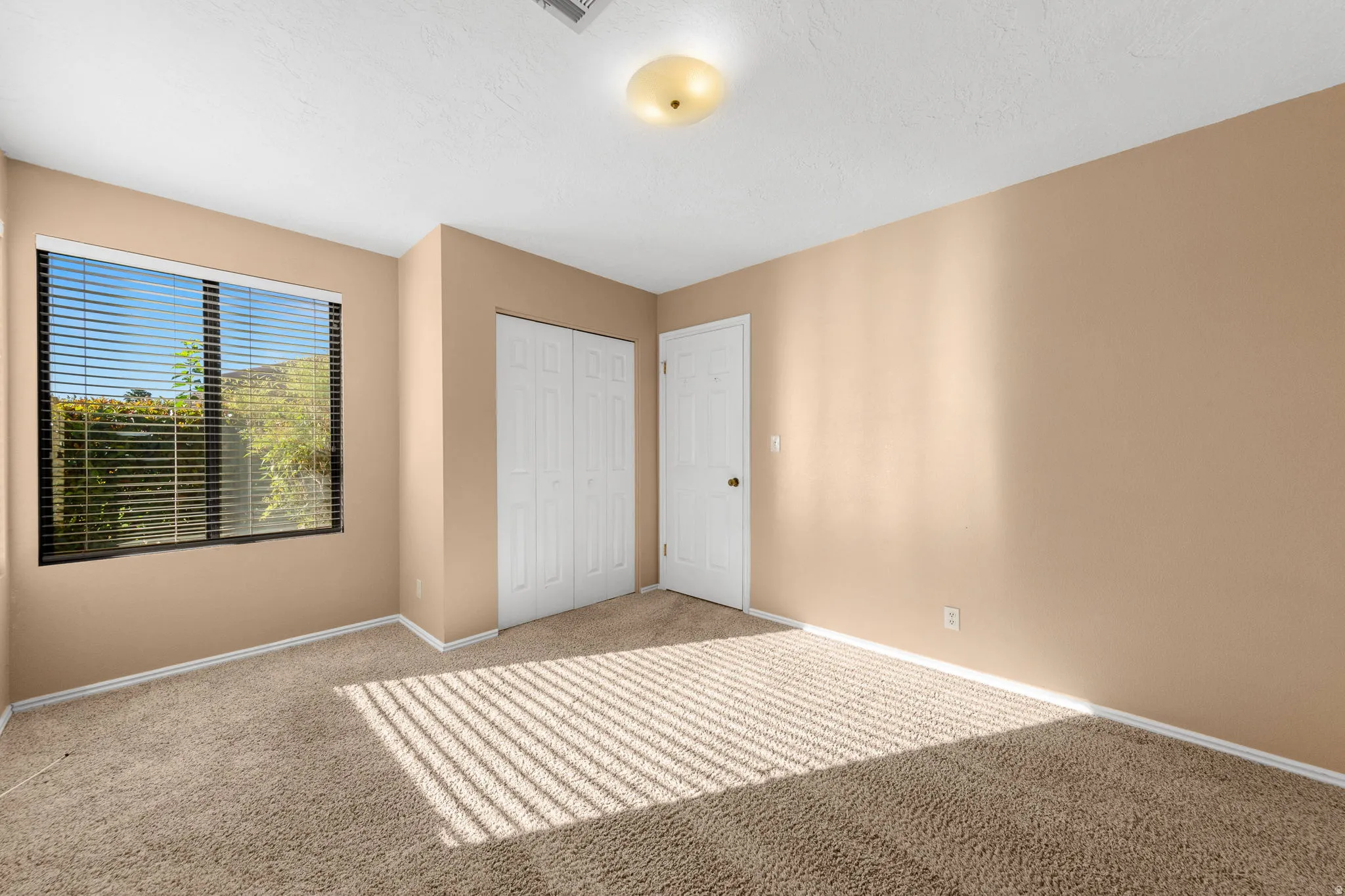 Bedroom 2. Neutral-toned wall paint with a window featuring horizontal blinds, textured carpet flooring, and a flush mount ceiling light