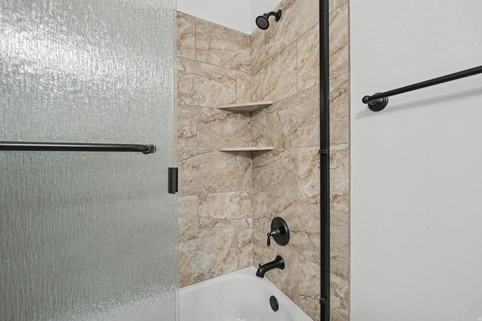 Bathtub shower with textured glass enclosure, tiled surround, built-in corner shelves, and matte black fixtures